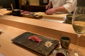 A sushi chef dressed in a clean white uniform works behind a polished wooden counter, expertly slicing fish with a sharp knife on a light wooden cutting board. In the foreground, a dark, textured ceramic platter holds a single piece of pink tuna nigiri sushi topped with a small relish, accompanied by a flower-shaped mound of pickled ginger. To the right of the plate rests a small ceramic cup, a dish of soy sauce, and a stemmed wine glass filled with a light, golden beverage under warm, intimate ambient lighting.