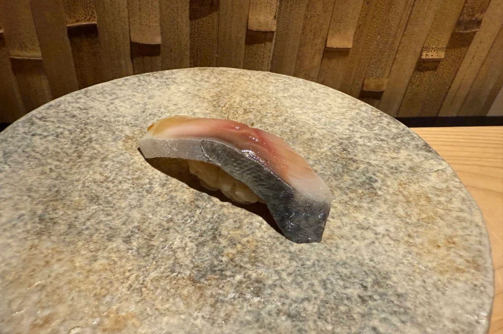 A single piece of mackerel nigiri sushi rests gracefully atop a large, circular stone serving plate with a mottled, rustic beige texture. The slice of fish features a glistening silver-and-blue skin doused in a light glaze, draped over a small, hand-pressed mound of seasoned sushi rice against a backdrop of a warm, textured bamboo wall panel.