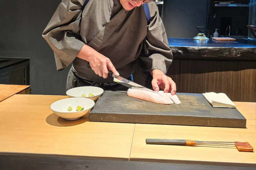 A chef dressed in a traditional grey robe uses a long, sharp knife to slice pale pink fish fillets on a dark slate board set atop a light wooden counter. Small white dishes with citrus lime wedges sit nearby, alongside metal long-handled cooking utensils arranged on the counter surface.