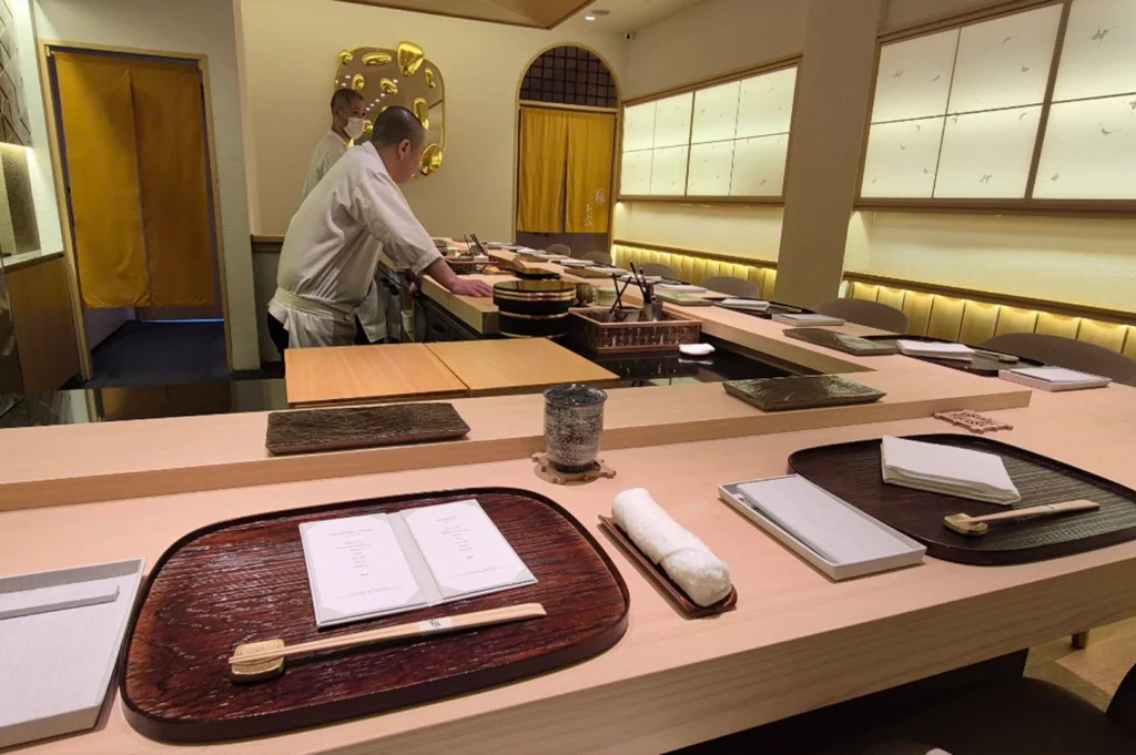 An eye-level view looks down the length of a polished wooden omakase counter set with individual placemats, wooden trays, folded white hand towels, and chopsticks. Behind the counter, two sushi chefs wearing white chef coats and face masks prepare ingredients near traditional sliding shoji screen walls illuminated by soft ambient light.