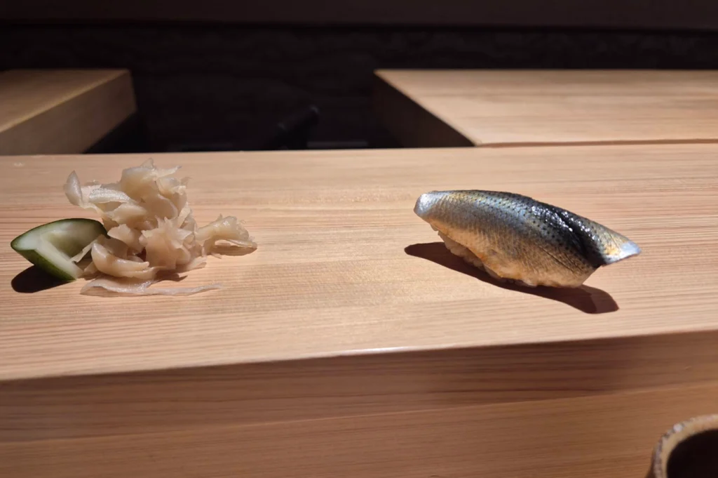 A single piece of nigiri sushi featuring silvery, patterned fish skin (kohada/gizzard shad) meticulously sliced and placed over sushi rice rests on a smooth light-wood counter. To its left sits a mound of pale pickled ginger alongside a small cucumber garnish, casting soft shadows across the clean wooden surface.