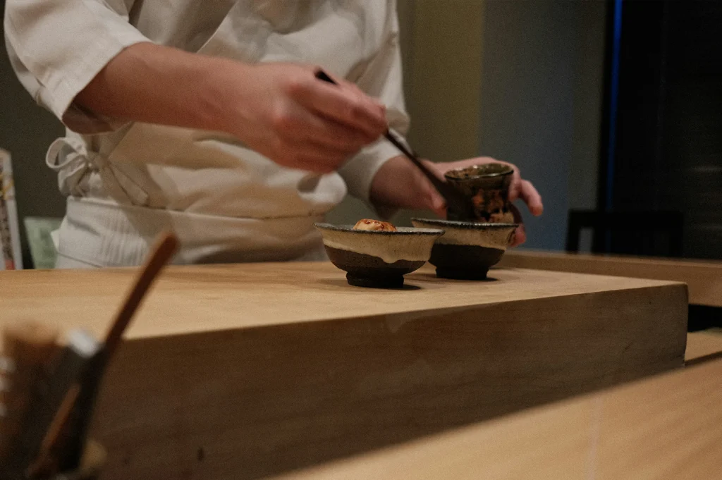 An eye-level shot captures a sushi chef wearing a white chef's coat working behind a smooth light-wood omakase counter. The chef uses fine wooden chopsticks to carefully place an ingredient into one of two small, rustic earthenware dishes sitting on the wooden serving block.