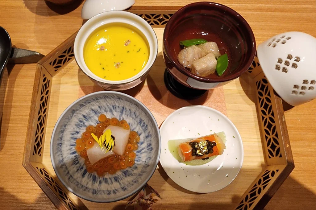 A high-angle shot of a multi-course appetizers served inside a hexagonal wooden tray, including a small bowl of yellow soup, a dark bowl with broth and vegetables, a blue-and-white dish with raw fish and salmon roe (ikura), and a white plate presenting a small seafood roll garnished with gold leaf.