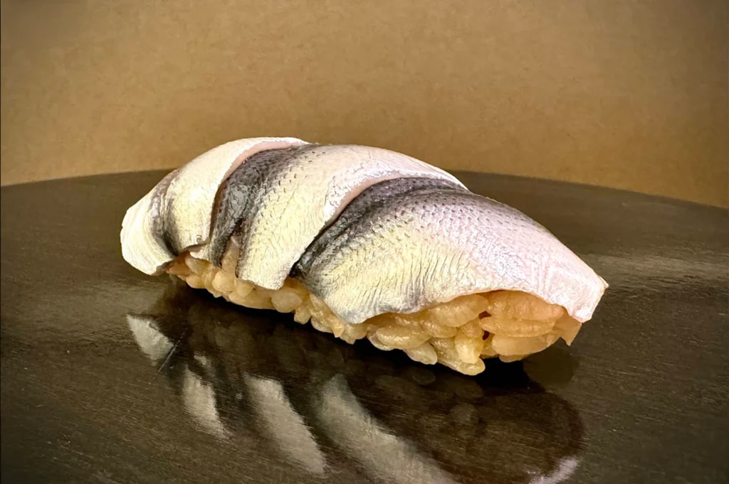 A close-up studio photograph presents a piece of kohada (gizzard shad) nigiri sushi resting on a dark reflective surface. The shiny, silvery fish skin displays delicate diagonal scoring along its back, draped gracefully over seasoned akazu (red vinegar) sushi rice.