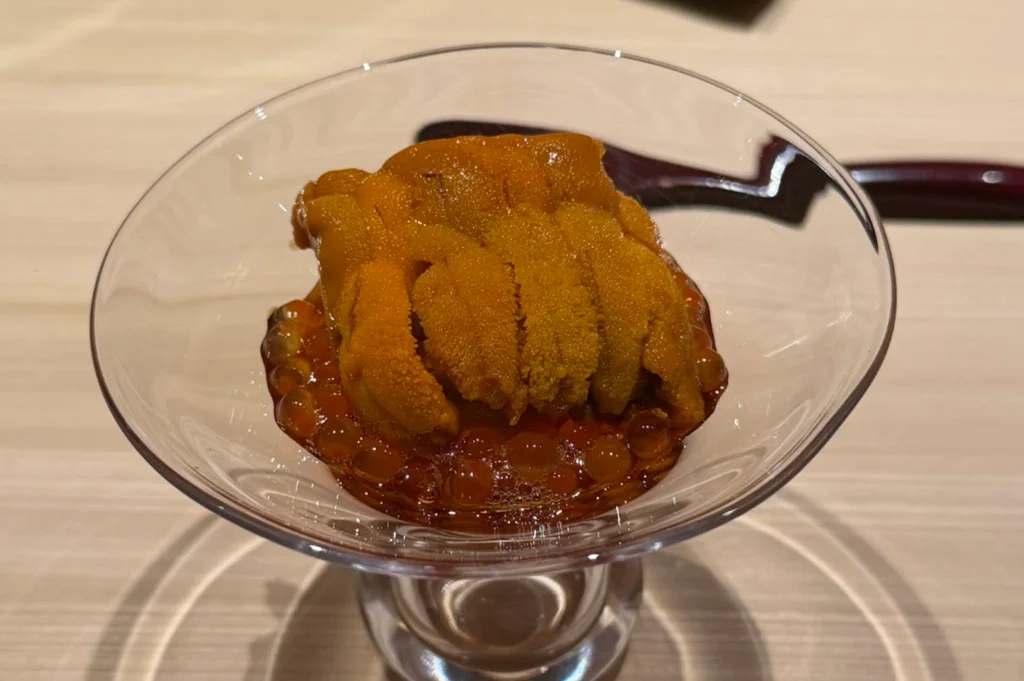 A close-up shot of a clear glass footed dish filled with a layer of glistening, amber-colored salmon roe (ikura) topped with a rich, textured mound of bright yellow-orange sea urchin (uni). In the soft-focused background, a dark wooden spoon rests on a light wooden table counter.