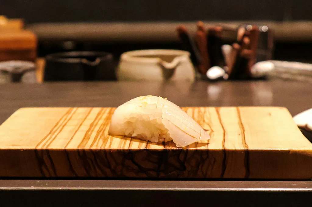 A high-resolution shot highlights a single piece of squid nigiri served on a rectangular olive wood board displaying visible natural wood grain patterns. The translucent white squid is precisely scored across its surface and lightly garnished with citrus zest, with ceramic condiment jars softly blurred in the background.