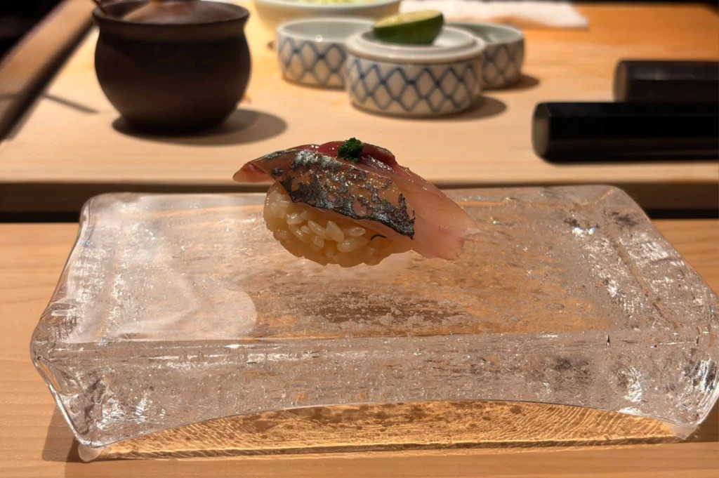 A close-up view shows a small mound of raw, uncooked short-grain white rice heaped on a speckled beige stone countertop, accompanied by two dried stalks of rice grain resting across the front. In the soft-focus background, a large white container and wooden kitchen items sit near the edge of a stainless steel sink, highlighting the pure ingredients used in sushi making.