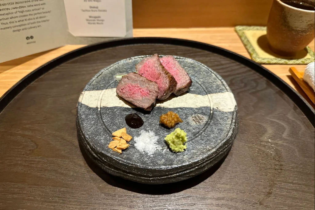 Three thick slices of rare, pan-seared beef showcasing bright pink centers are served on a round, textured grey ceramic plate. Below the meat, an array of colorful condiments—including wasabi, sea salt, fried garlic chips, and savory pastes—are neatly arranged along the bottom edge.
