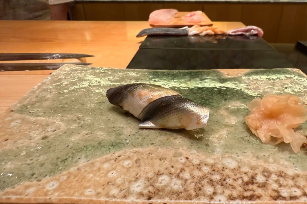A detailed shot features a piece of pickled gizzard shad nigiri placed in the center of a rectangular speckled green-and-tan ceramic serving plate. A small cluster of thinly sliced pickled ginger sits on the right side of the plate, with the chef's wooden prep station and prep knives faintly visible in the blurred background.