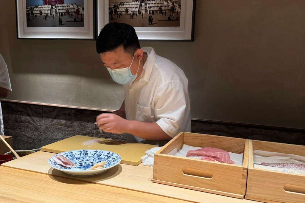 A sushi chef wearing a white short-sleeved uniform shirt and a blue protective face mask leans over a light wooden counter while carefully preparing food. In front of him sits a blue-and-white patterned ceramic plate and wooden preparation boxes containing fresh cuts of fish, with two framed black-and-white photographs hanging on the wall behind him.