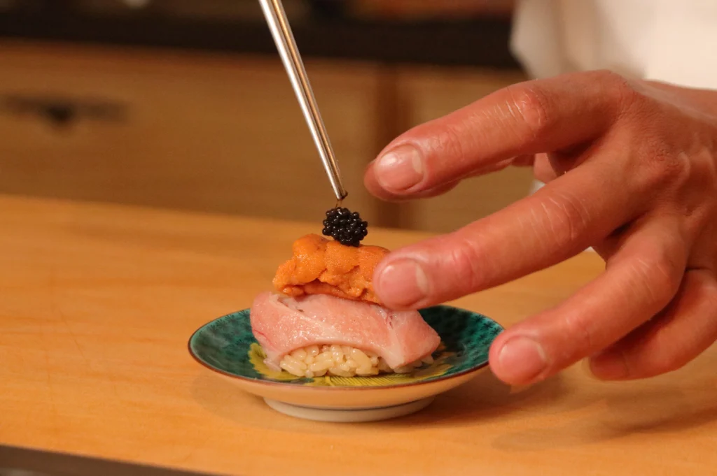 A close-up photograph shows a chef using long metal tweezers to place a cluster of dark black caviar atop a piece of uni (sea urchin) and fatty tuna nigiri. The decadent sushi assembly rests on a small, patterned blue-and-yellow ceramic plate set against a warm, light wooden prep surface.