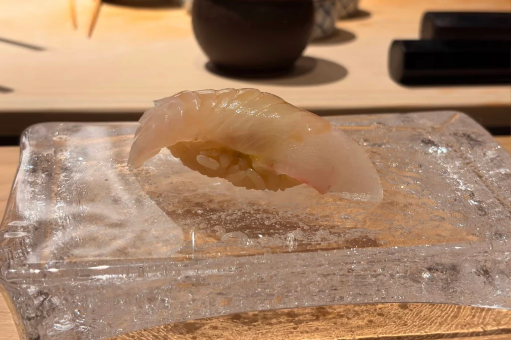 A piece of translucent, pale white fish nigiri sushi with delicate scoring is presented on a thick, textured clear glass platter that sits on a warm wooden counter. The fish has a glossy finish and rests over a compact bed of seasoned sushi rice, while the blurred background reveals traditional teacups and small ceramic tableware.