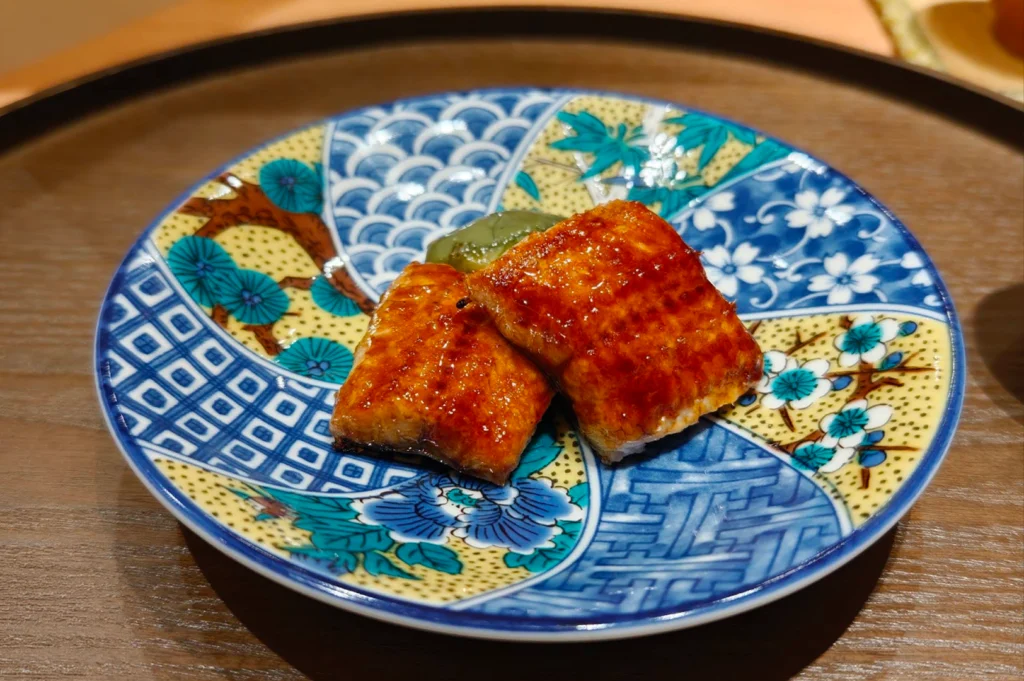 Two pieces of glazed, grilled unagi (freshwater eel) featuring a glossy golden-brown finish rest on a vibrant, multi-colored Japanese ceramic plate decorated with traditional blue, green, and yellow patterns.