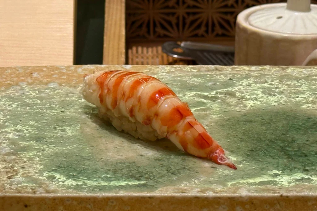 A close-up photograph captures a piece of kuruma ebi (Japanese tiger prawn) nigiri served on a textured, glazed ceramic platter. The cooked prawn shows vibrant orange and white striped bands over seasoned sushi rice, with a traditional ceramic soy pourer set in the background.