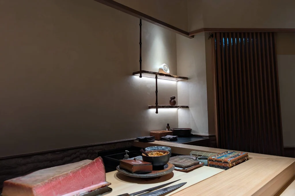A long wooden sushi bar counter featuring a large block of fresh pink tuna, a sharp chef's knife, small ceramic bowls, and traditional preparation tools. Illuminated floating shelves with subtle warm backlighting display small pottery items against neutral walls in the background, set alongside dark vertical slatted wall panels.