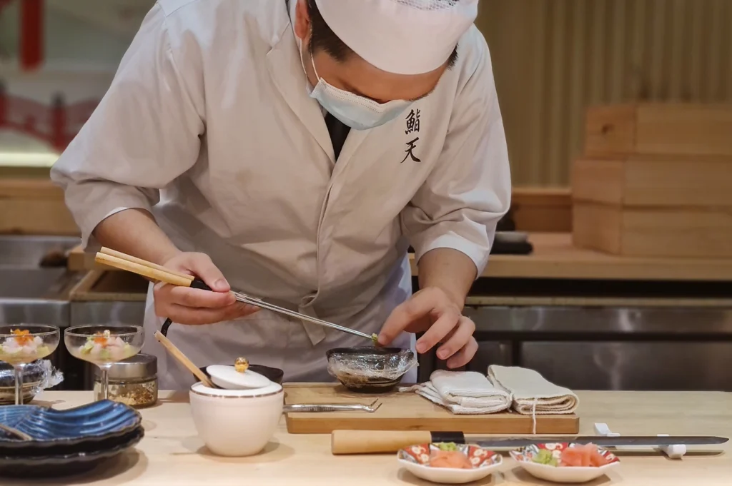 A sushi chef wearing a white uniform, face mask, and chef's hat leans over a wooden prep station to delicately arrange garnishes with chopsticks. Arranged along the clean counter are small dishes of pickled ginger, a white sauce jar, a long yanagiba sushi knife, and two stemless cocktail glasses containing layered appetizers.