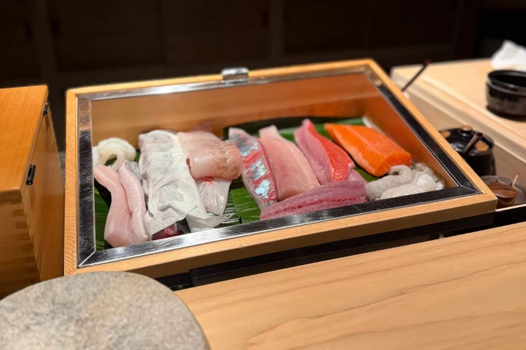 A wooden display box with a clear glass lid sits open on a sushi counter, showcasing a pristine arrangement of fresh, raw seafood fillets resting on green banana leaves. The colorful selection includes bright orange salmon, deep red tuna, silver-skinned mackerel, and pale white fish fillets, lined up neatly alongside other sushi preparation tools and wooden containers.