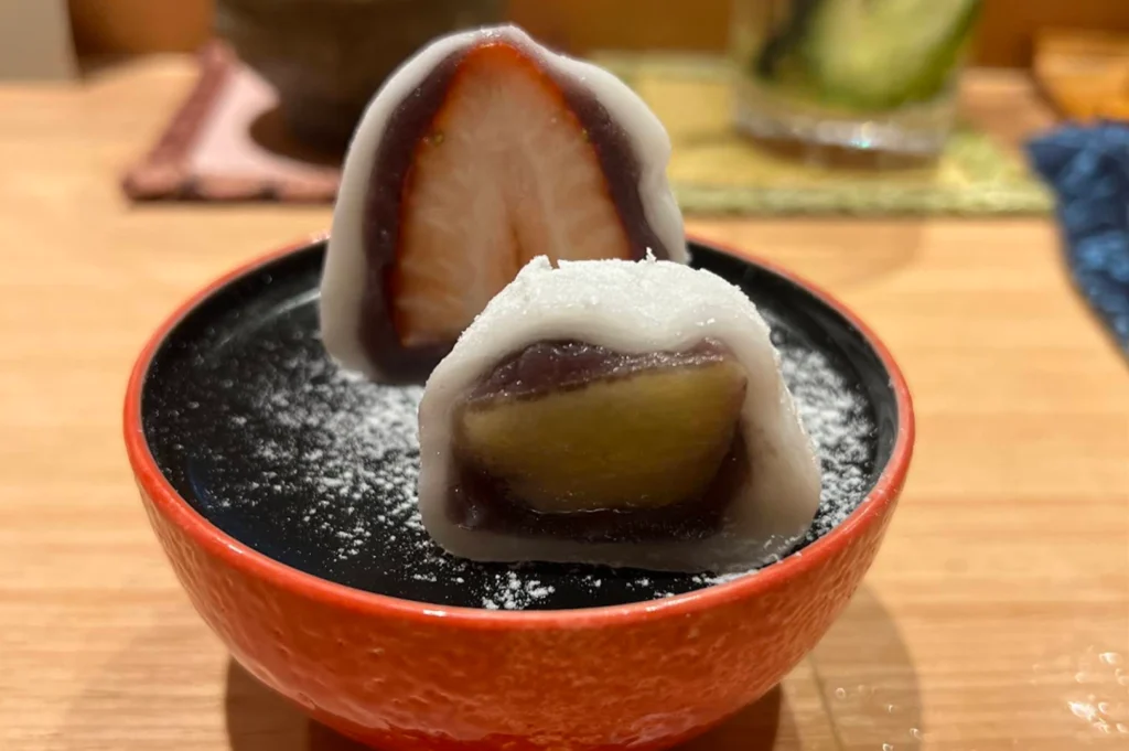 A red ceramic dish holds two halved mochi desserts lightly dusted with powdered sugar, displaying a whole fresh strawberry encased in sweet red bean paste inside one piece and a fruit filling in the other.