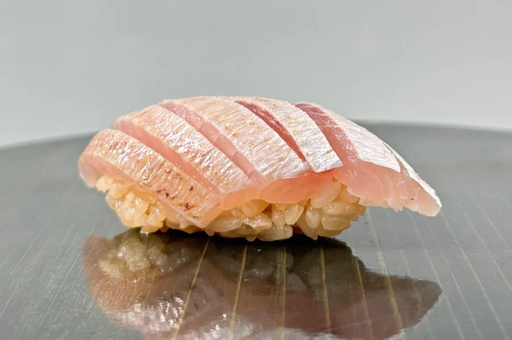 A macro profile shot highlights a piece of kasugodai (young sea bream) nigiri resting on a dark, wood-grained lacquered surface. The translucent, pinkish fish fillet is precisely scored in parallel lines to reveal the delicate flesh beneath, sitting atop reddish-hued seasoned rice.