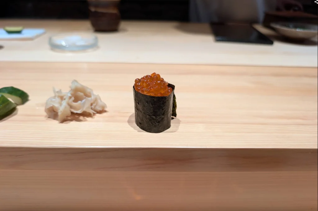 A single piece of ikura (salmon roe) gunkan sushi sits upright on a light-colored wooden sushi counter, wrapped in crisp dark nori seaweed and generously topped with glistening, translucent orange salmon eggs. To the left, a delicate heap of thinly sliced pickled ginger (gari) and a cucumber slice rest on the smooth wooden surface, set against a softly blurred background of the dining space.
