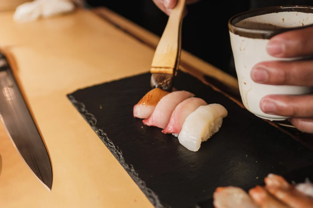 An overhead view shows a chef holding a small sauce cup and using a wide wooden brush to apply soy glaze over four pieces of assorted nigiri sushi. The nigiri pieces—including tuna, yellowtail, and white fish—are arranged neatly on a rectangular black slate tray resting on a light-colored wooden table next to a sharp chef's knife.