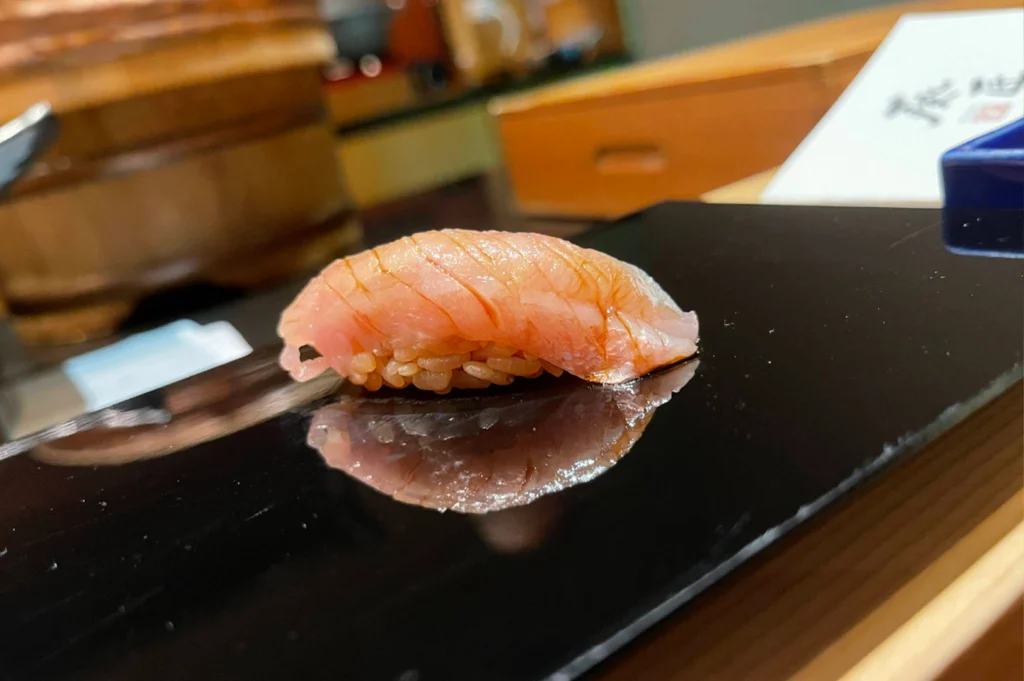 A single piece of fatty tuna nigiri sushi, scored with fine parallel cuts, sits on a reflective black serving platter that creates a clear mirror-like reflection of the sushi on its polished surface.